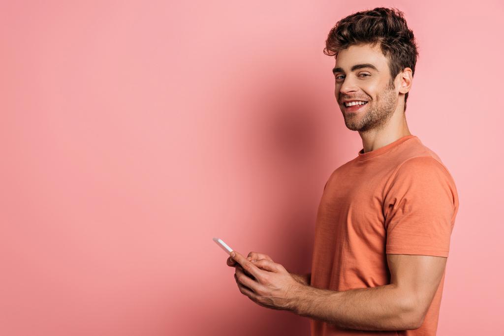 cheerful young man smiling at camera while chatting on smartphone on pink background - 写真・画像