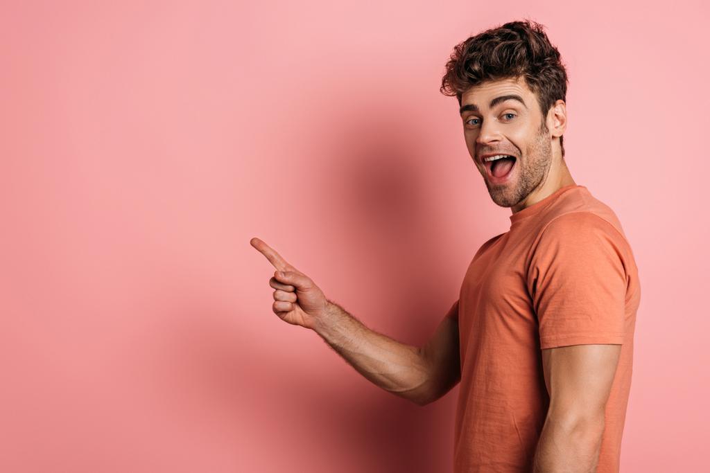 vzrušený mladý muž ukazuje prstem při pohledu na kameru na růžovém pozadí - Fotografie, Obrázek