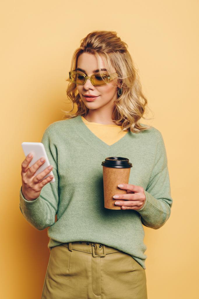 стильная, улыбчивая девушка, держащая в руках кофе, пока она разговаривает по смартфону на желтом фоне
 - Фото, изображение
