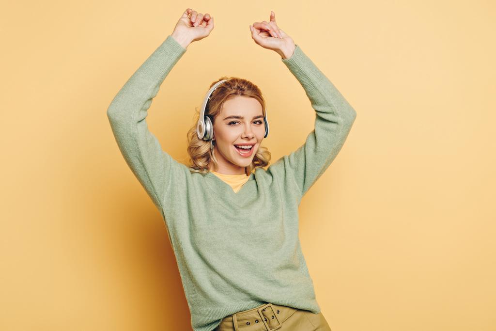 cheerful girl listening music in wireless headphones, dancing and looking at camera on yellow background - Photo, Image