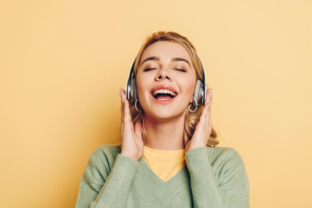 счастливая девушка слушает музыку в беспроводных наушниках с закрытыми глазами на желтом фоне
 - Фото, изображение