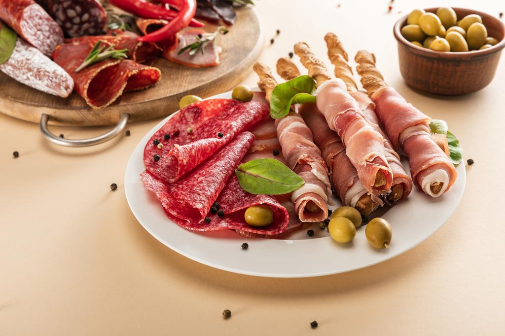 смачні м'ясні тарілки, подані з оливками, спеціями та паличками на тарілці та дерев'яною дошкою на бежевому фоні
 - Фото, зображення