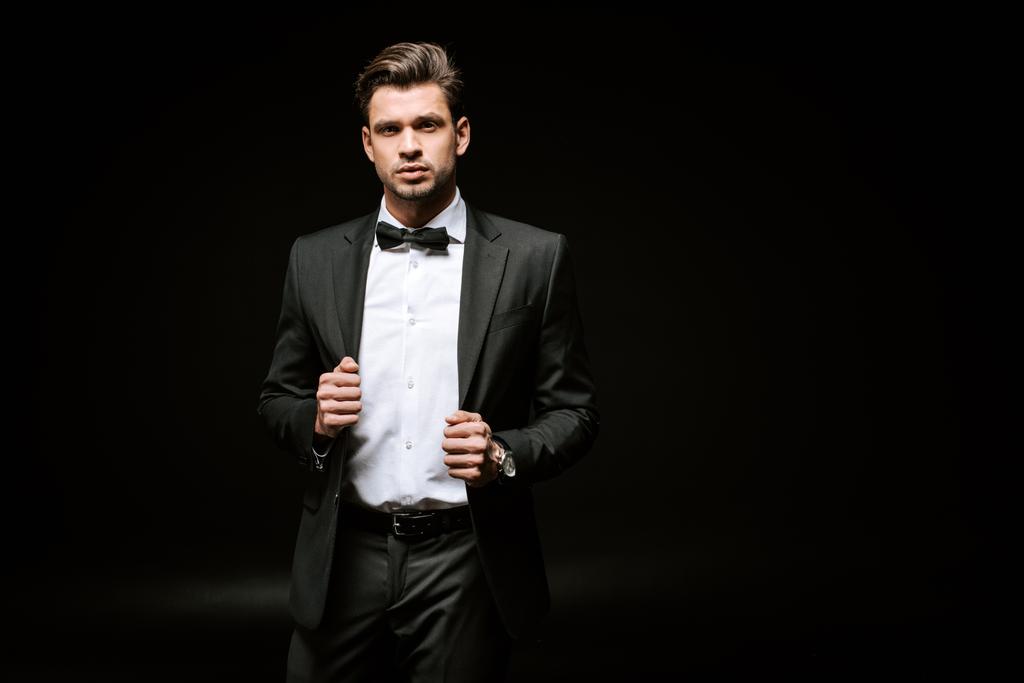 Eleganter Mann im Anzug blickt in die Kamera und berührt Blazer auf Schwarz  - Foto, Bild