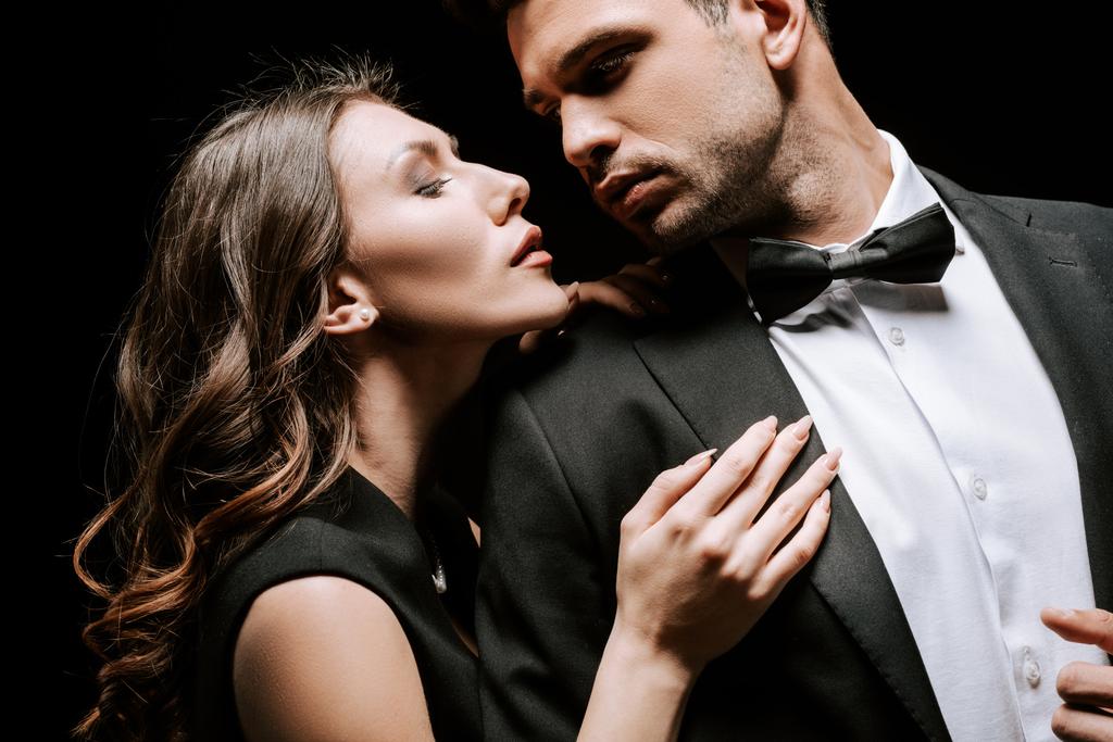 νεαρή ελκυστική γυναίκα που αγγίζει όμορφο άνδρα με κοστούμι απομονωμένο σε μαύρο  - Φωτογραφία, εικόνα