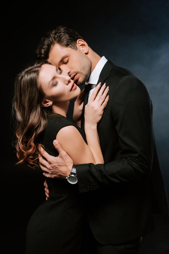 όμορφος άντρας αγκαλιάζει ελκυστική γυναίκα σε μαύρο με καπνό  - Φωτογραφία, εικόνα
