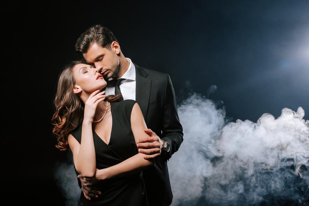 knappe man in pak knuffelen aantrekkelijke vrouw op zwart met rook  - Foto, afbeelding