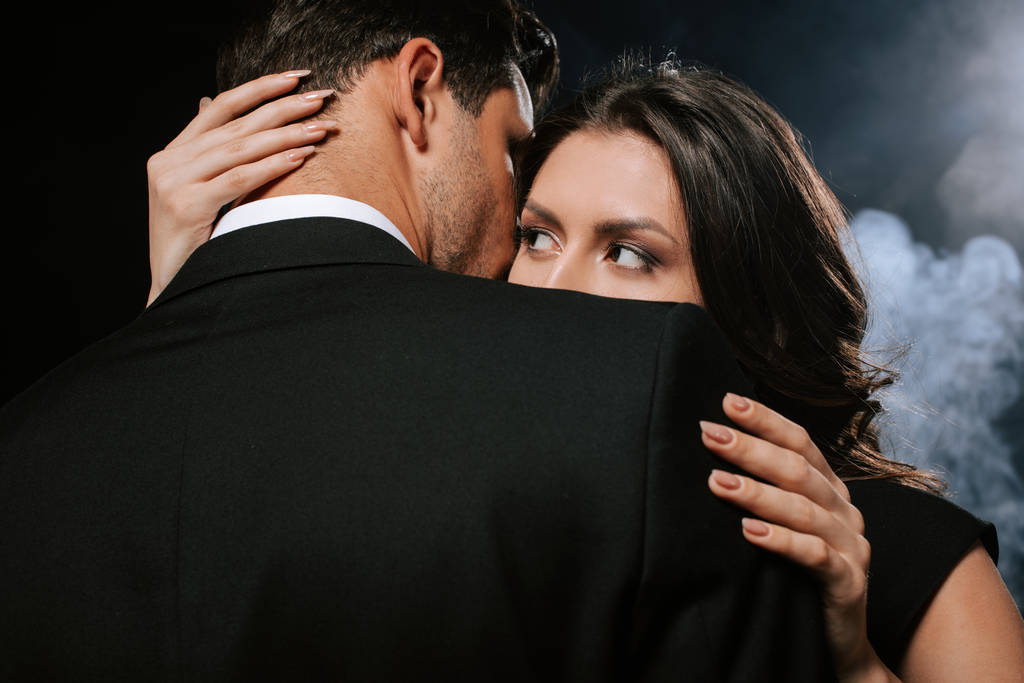 jong vrouw kijken naar man terwijl knuffelen op zwart met rook  - Foto, afbeelding