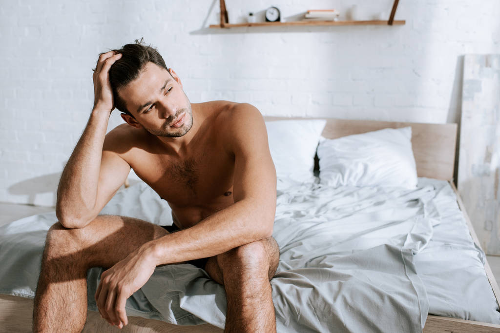 muskularny mężczyzna odwracając wzrok siedząc na łóżku i dotykając włosów  - Zdjęcie, obraz