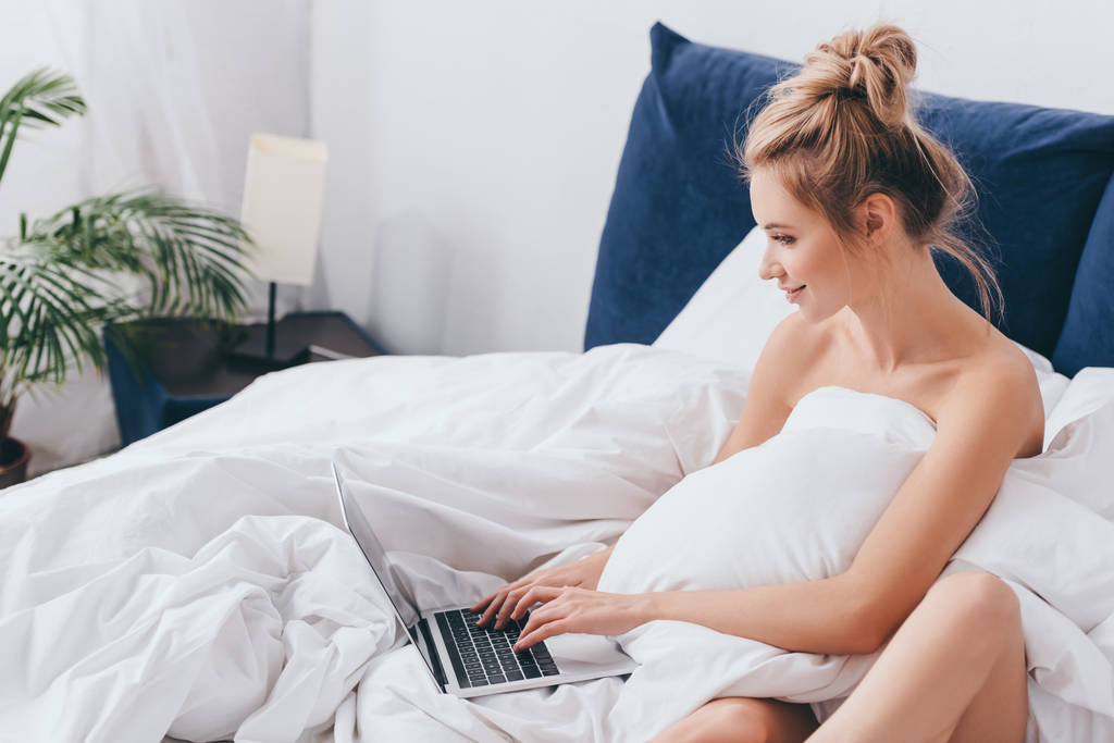 χαμογελαστή γυναίκα που χρησιμοποιεί φορητό υπολογιστή στο κρεβάτι με σεντόνια το πρωί  - Φωτογραφία, εικόνα