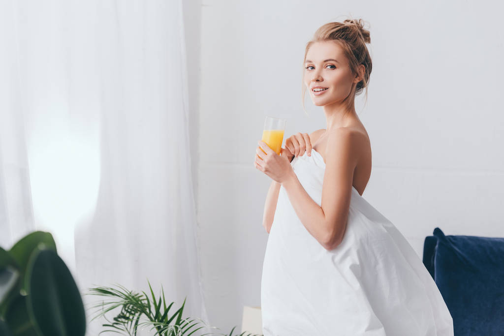 mujer sonriente con vaso de jugo en sábanas blancas en el dormitorio por la mañana
 - Foto, Imagen