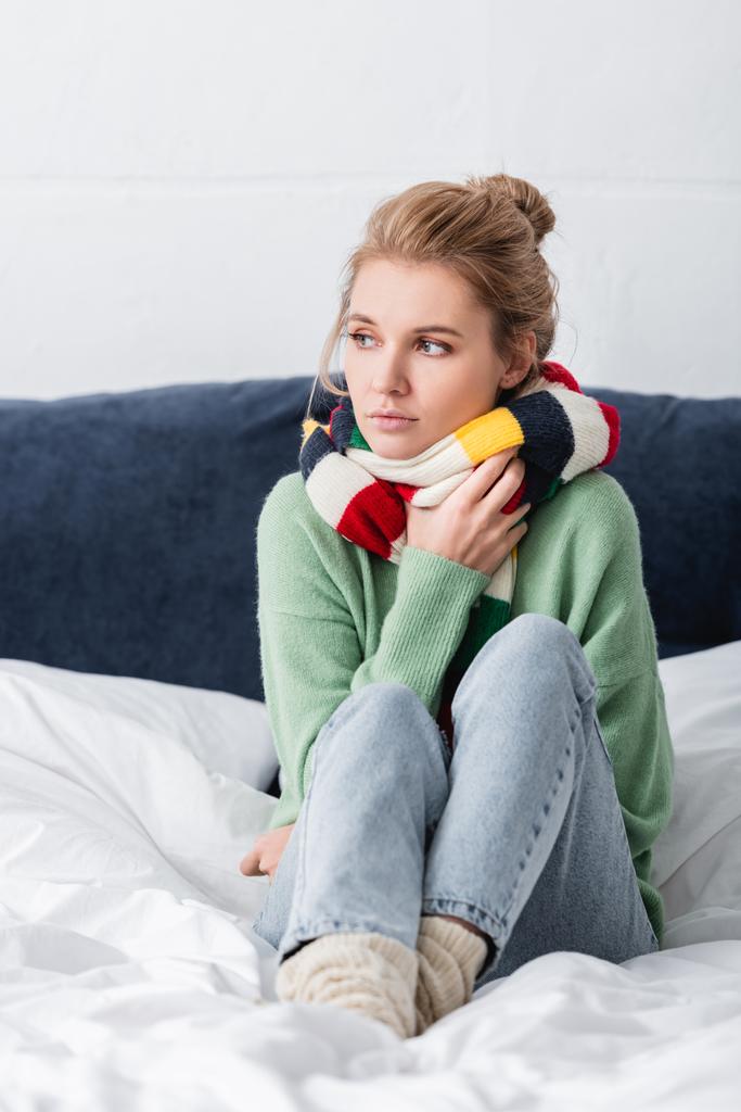 молодая больная женщина в свитере и шарфе сидит на кровати
  - Фото, изображение