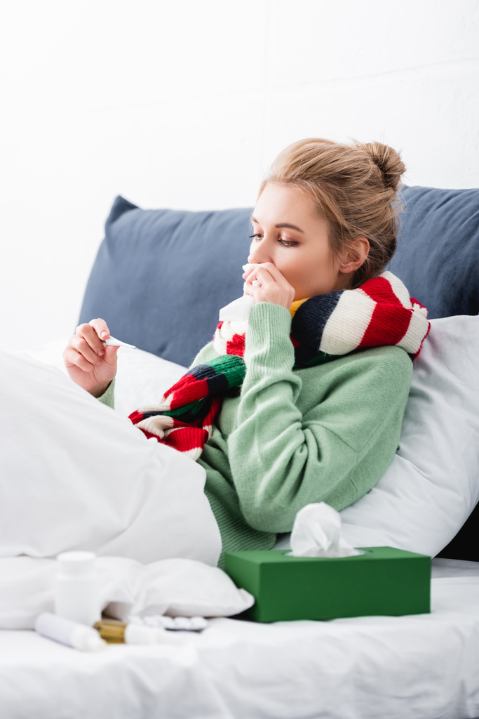 ill woman in scarf having fever and looking at thermometer in bed  - Photo, Image