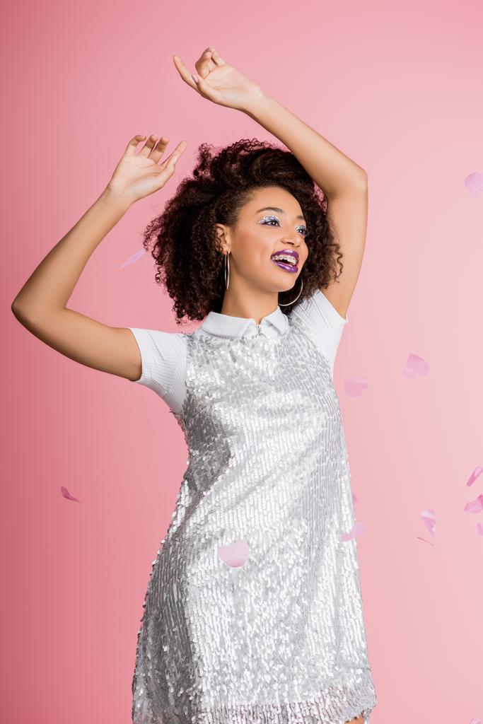 attractive happy african american girl with dental braces, with silver glitter eyeshadows and purple lips dancing in paillettes dress, isolated on pink with confetti - Photo, Image