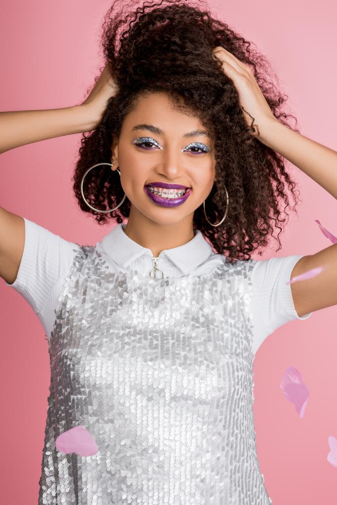 cheerful african american girl with dental braces, with silver glitter eyeshadows and purple lips wearing paillettes dress, isolated on pink with confetti - Photo, Image