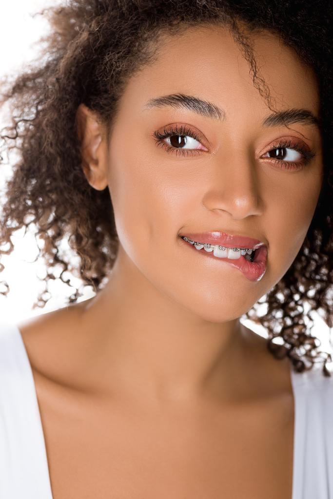 Χαρούμενη Αφροαμερικανή με οδοντικά σιδεράκια που δαγκώνουν χείλια, απομονωμένη στα λευκά - Φωτογραφία, εικόνα
