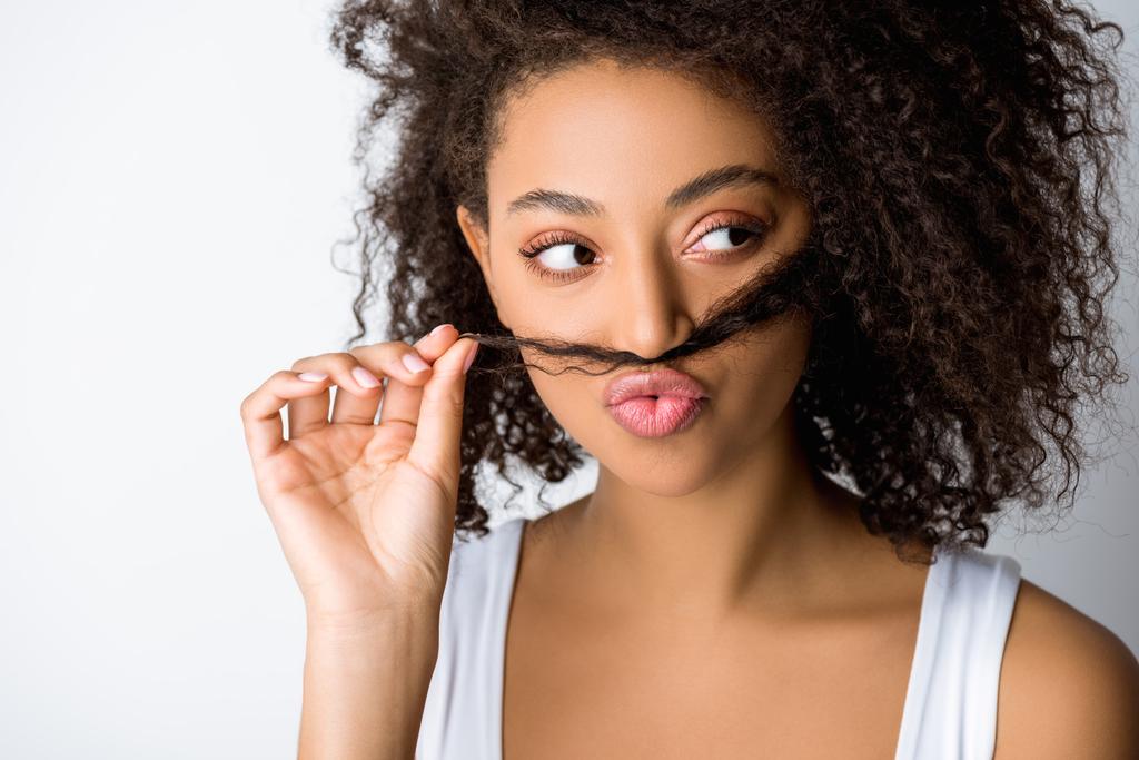 αστεία ελκυστική Αφροαμερικανή που φτιάχνει ψεύτικο μουστάκι από μαλλιά, απομονωμένη σε γκρι. - Φωτογραφία, εικόνα