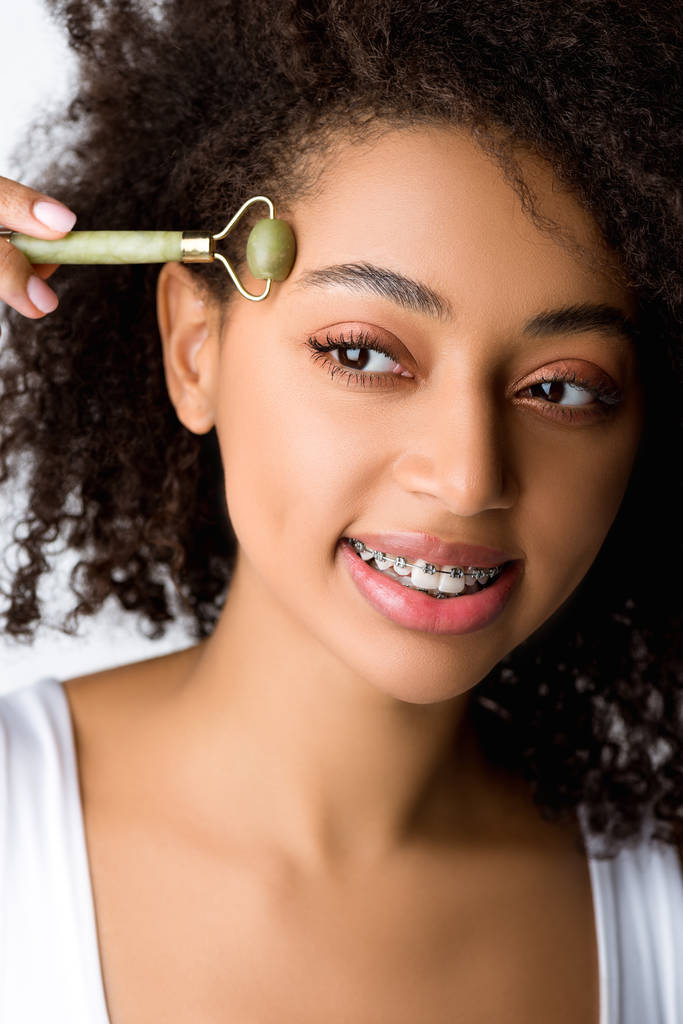 bela menina afro-americana sorridente com aparelho dentário usando ferramenta de massagem facial, isolado em cinza
  - Foto, Imagem