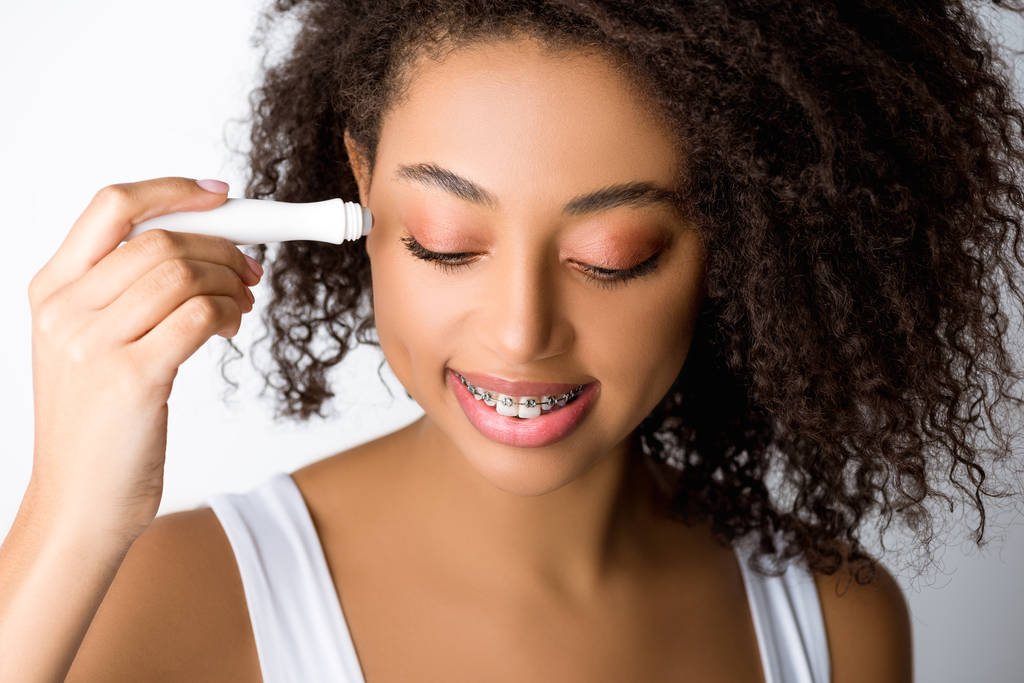 sorrindo menina afro-americana com aparelho dentário usando rolo olho hidratante, isolado em cinza
 - Foto, Imagem