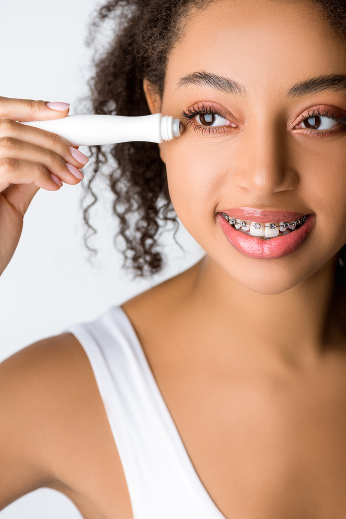 šťastná africká americká žena se zubními rovnátky pomocí hydratačního očního válce, izolované na šedé - Fotografie, Obrázek
