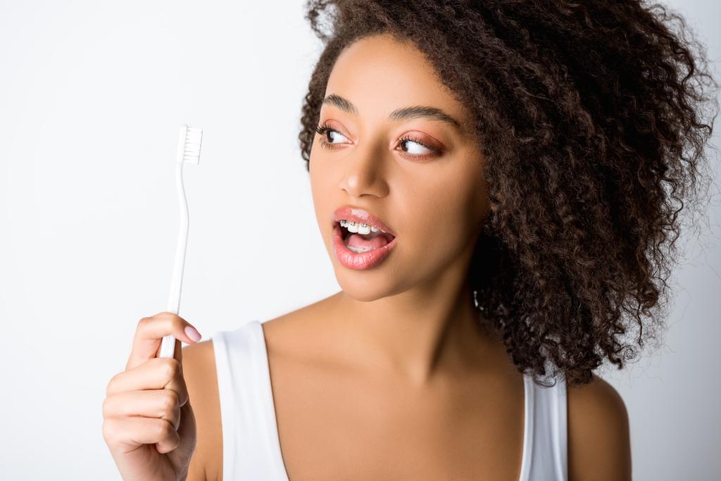 femme afro-américaine surprise avec des appareils dentaires regardant la brosse à dents, isolé sur gris
 - Photo, image