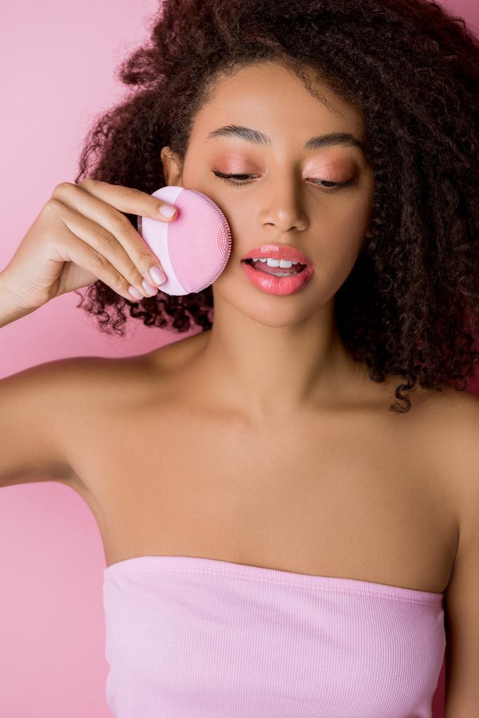 Αφροαμερικανή γυναίκα με κλειστά μάτια που χρησιμοποιεί βούρτσα προσώπου καθαρισμού σιλικόνης, απομονωμένη σε ροζ - Φωτογραφία, εικόνα