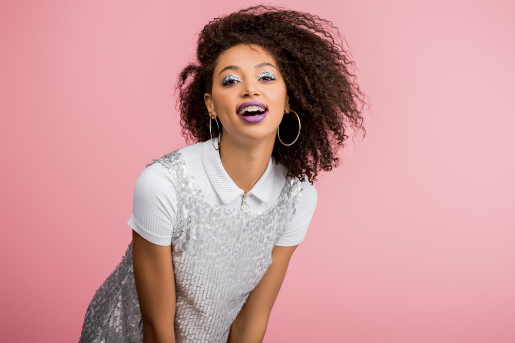 élégante fille afro-américaine souriante avec des bretelles dentaires, avec des fards à paupières à paillettes argentées et des lèvres violettes dansant en robe paillettes, isolée sur rose
 - Photo, image