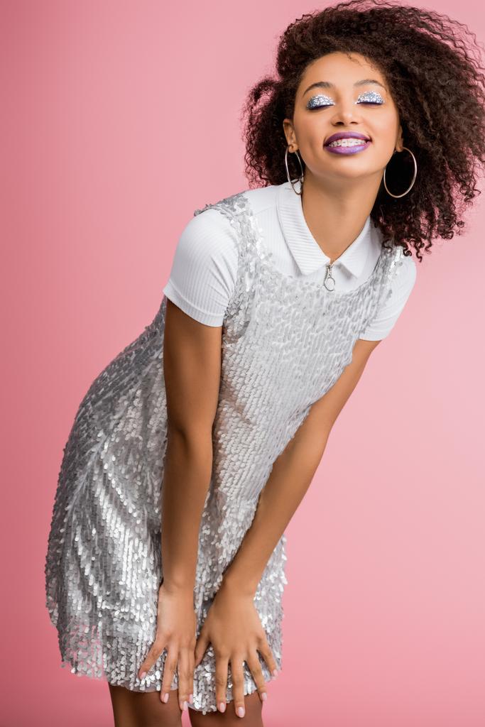 mosolygós afro-amerikai lány fogszabályzóval, ezüst csillogó szemhéjjal és lila ajkakkal táncoló paillette ruha, elszigetelt rózsaszín - Fotó, kép