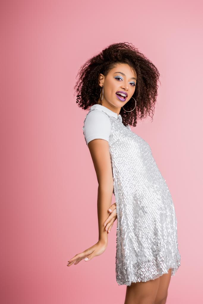 happy african american girl with dental braces, with silver glitter eyeshadows and purple lips dancing in paillettes dress, isolated on pink - Photo, Image