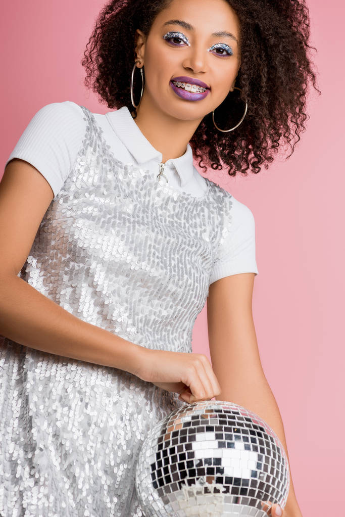 happy young african american woman in paillettes dress holding disco ball, isolated on pink  - Photo, Image