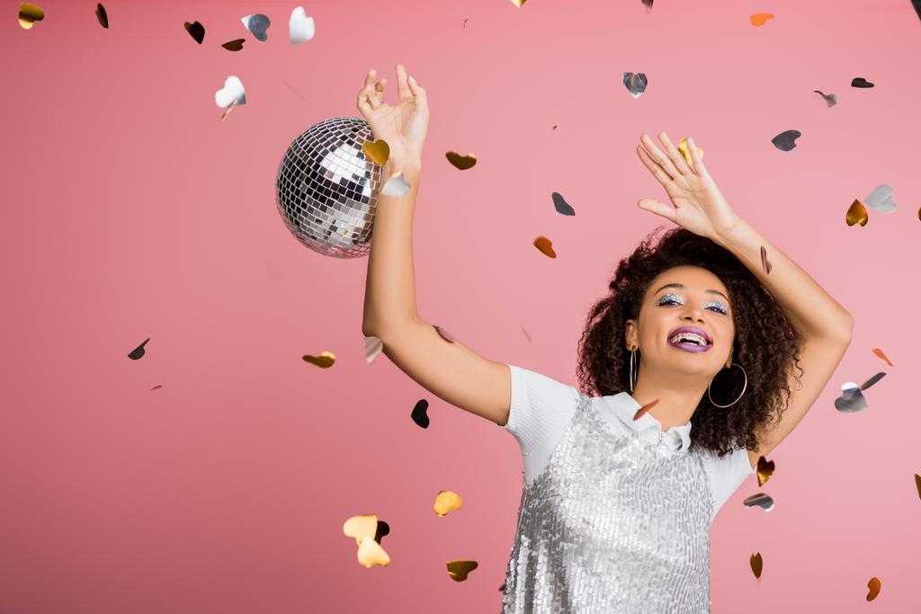 excited african american girl in paillettes dress dancing with disco ball, isolated on pink with confetti - 写真・画像