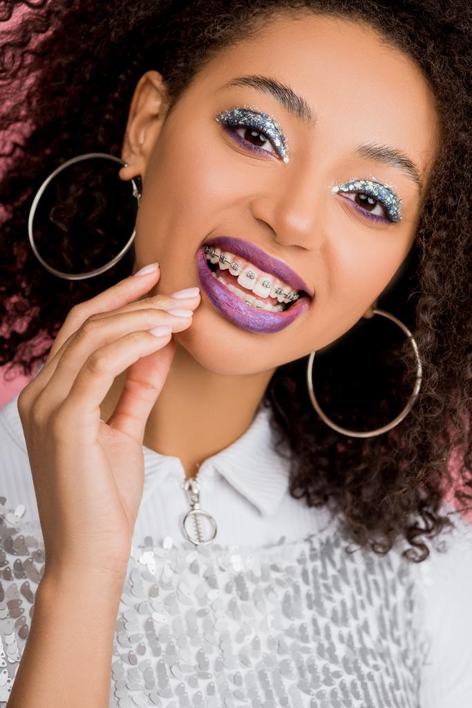 szczęśliwa Afroamerykanka dziewczyna z aparatem stomatologicznym, ze srebrnymi błyszczącymi oczkami i fioletowymi ustami w sukience w paillettes, odizolowana na różowo - Zdjęcie, obraz