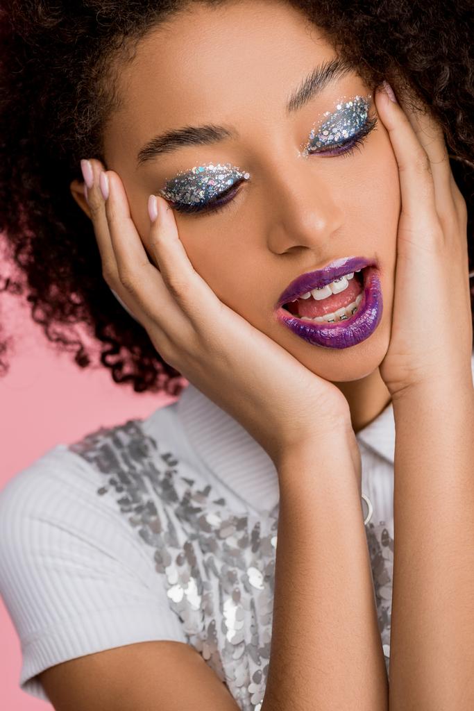 emotional african american girl with silver glitter eyeshadows and purple lips wearing paillettes dress, isolated on pink - 写真・画像