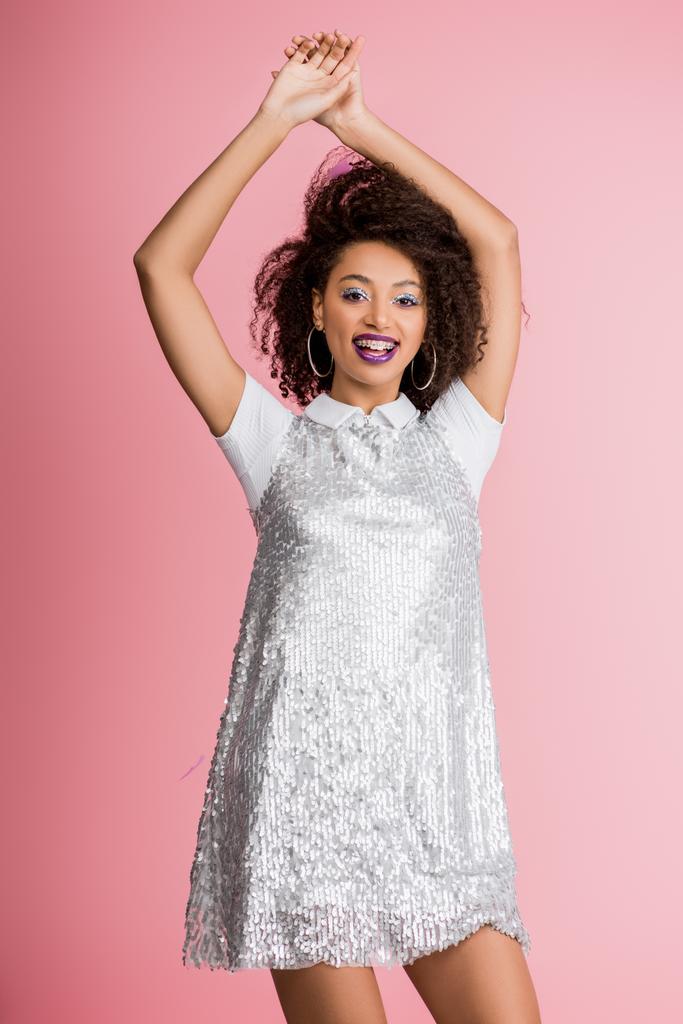 happy african american girl with silver glitter eyeshadows and purple lips dancing in paillettes dress, isolated on pink - Photo, Image