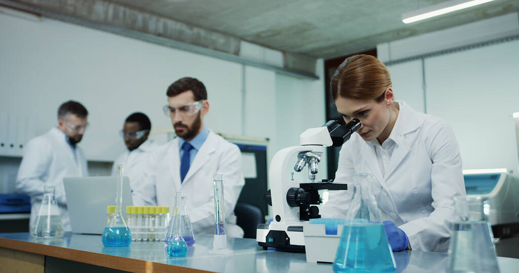 白人女性科学者で白いローブを着て顕微鏡を見ながらいくつかの調査と彼女の男性同僚テストいくつかの液体でチューブにラップトップコンピュータ. - 写真・画像