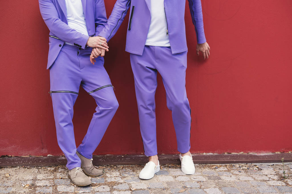 abgeschnittenes Bild eines homosexuellen Paares beim Händchenhalten. lila Anzüge rote Wand auf Hintergrund  - Foto, Bild