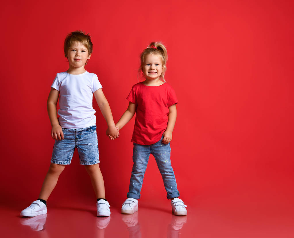 Small happy children boy and girl in stylish casual clothing standing, holding hands and feeling shy over red background - Photo, Image