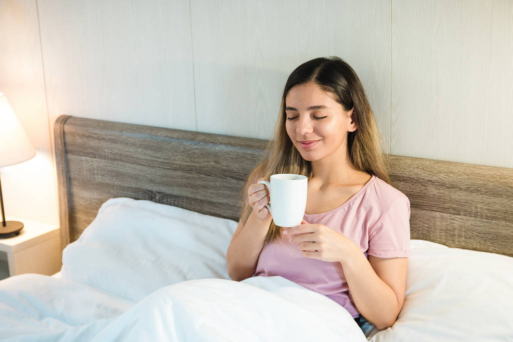 улыбающаяся красивая женщина с закрытыми глазами, держа чашку в постели с белыми постельными принадлежностями
 - Фото, изображение