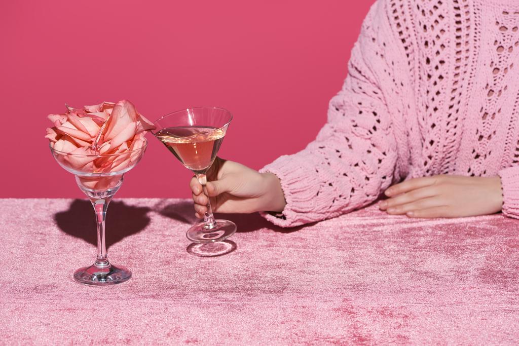 Kırpılmış bir kadın cam kenarında gül şarabı bardağı tutarken, kadife kumaş üzerinde gül yaprakları, pembe, kızsı bir konseptte izole edilmiş.  - Fotoğraf, Görsel