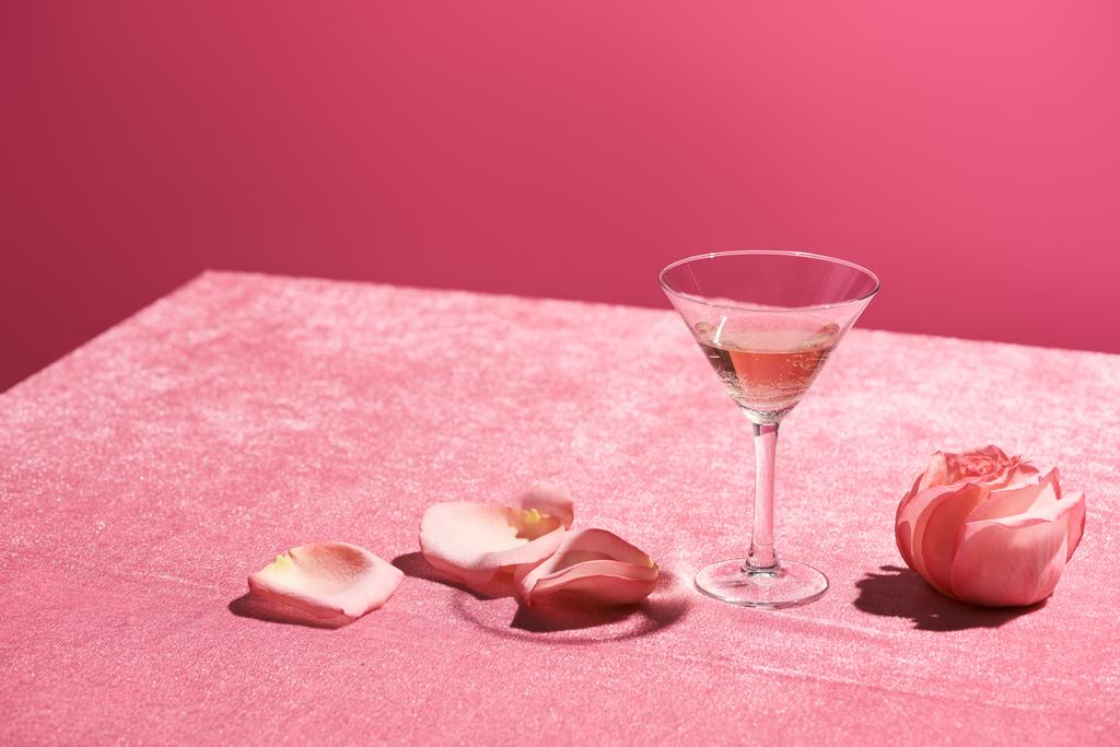 τριαντάφυλλο κρασί σε ποτήρι κοντά σε πέταλα και τριαντάφυλλο μπουμπούκι σε βελούδινο ροζ πανί που απομονώνονται σε ροζ, κοριτσίστικη έννοια - Φωτογραφία, εικόνα