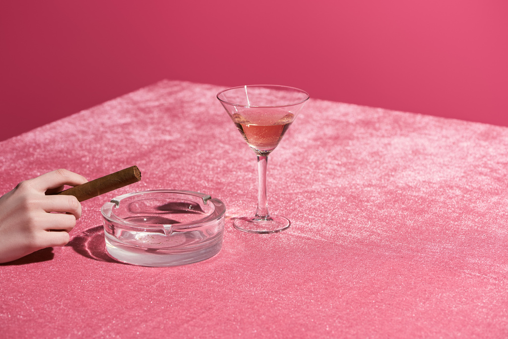 vista ritagliata della donna che tiene il sigaro vicino al posacenere e vetro di vino rosato su un panno di velluto isolato su rosa, concetto femminile
  - Foto, immagini