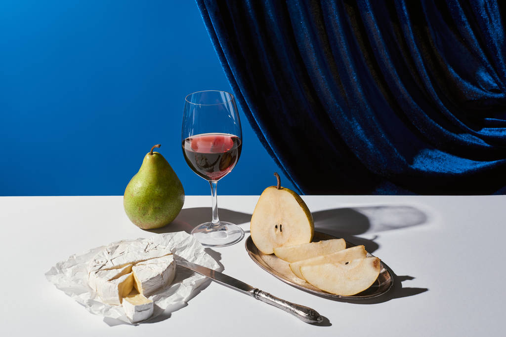 klasická zátiší s hruškami, červeným vínem a sýrem Camembert na bílém stole u sametové záclony izolované na modré - Fotografie, Obrázek