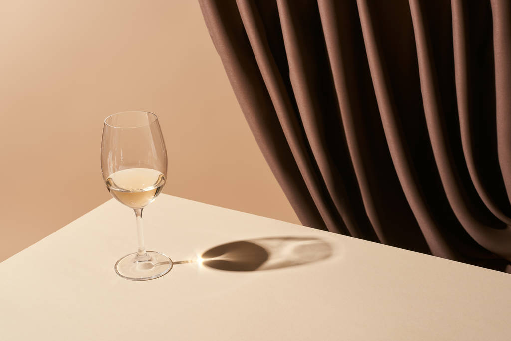 ベージュ色に隔離されたカーテンの近くのテーブルの上に白ワインのガラスと古典的な静物画 - 写真・画像