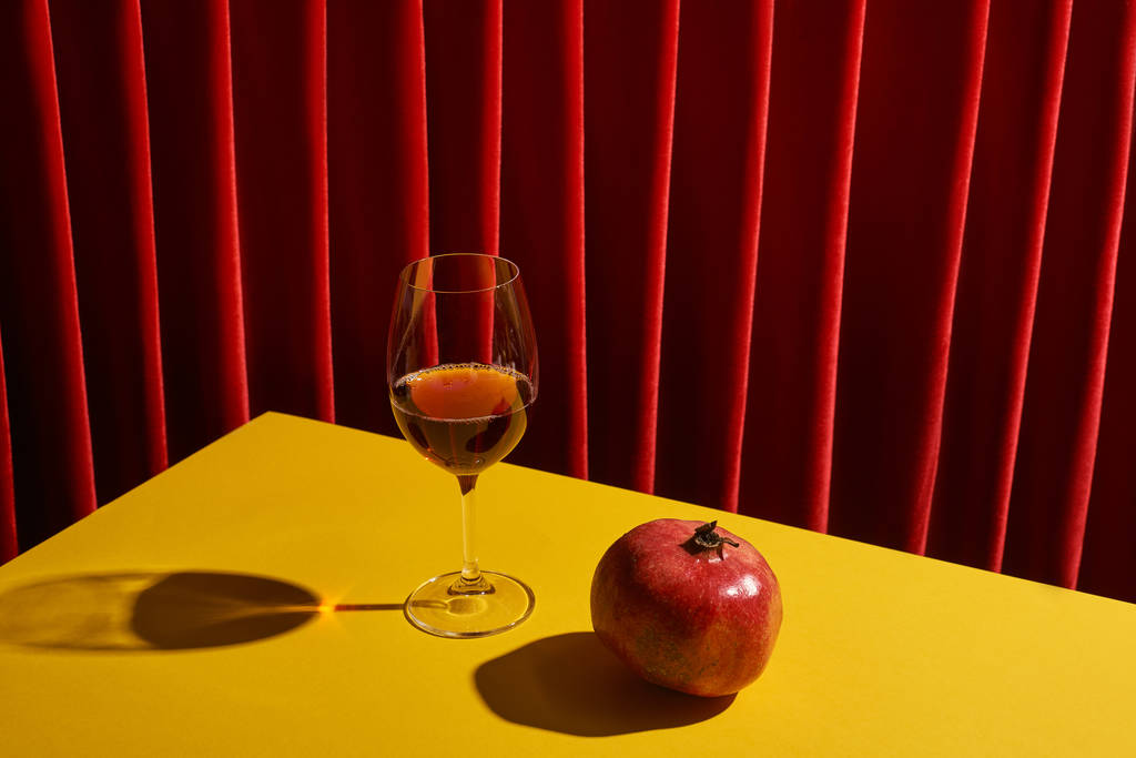klassieke stilleven met granaatappel in de buurt van glas rode wijn op gele tafel in de buurt van rood gordijn - Foto, afbeelding