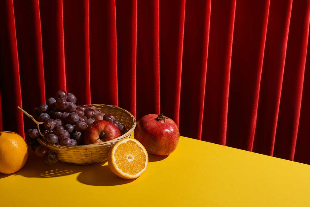 赤いカーテンの近くの黄色のテーブルの上にウィッカーバスケットの果物と古典的な静物画 - 写真・画像