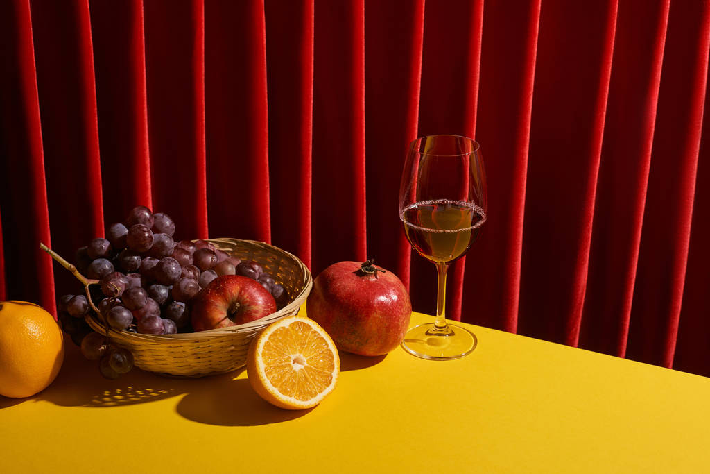 klassiek stilleven met fruit in rieten mandje bij glas rode wijn op gele tafel bij rood gordijn - Foto, afbeelding