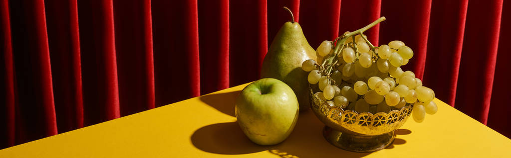 Kırmızı perde yakınındaki sarı masada yeşil meyveli klasik natürmort. - Fotoğraf, Görsel