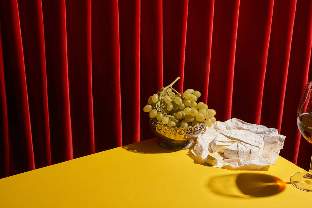 vida tranquila clássico com Camembert perto de copo de vinho tinto e uva na mesa amarela perto de cortina vermelha
 - Foto, Imagem