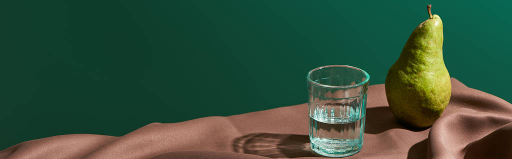 klasická zátiší s hruškou a vodou ve skle na stole s hnědým ubrusem izolovaným na zeleném panoramatickém záběru - Fotografie, Obrázek