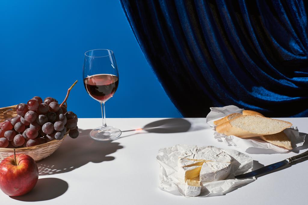 ブルーベローカーテン近くの白いテーブルの上に果物、赤ワイン、バゲット、キャンベルチーズと古典的な静物画 - 写真・画像
