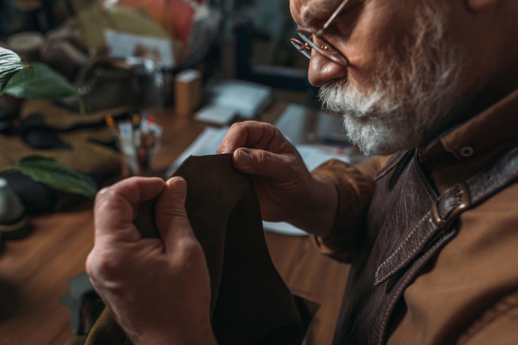 atento, zapatero senior sosteniendo pieza de cuero genuino en el taller
 - Foto, Imagen